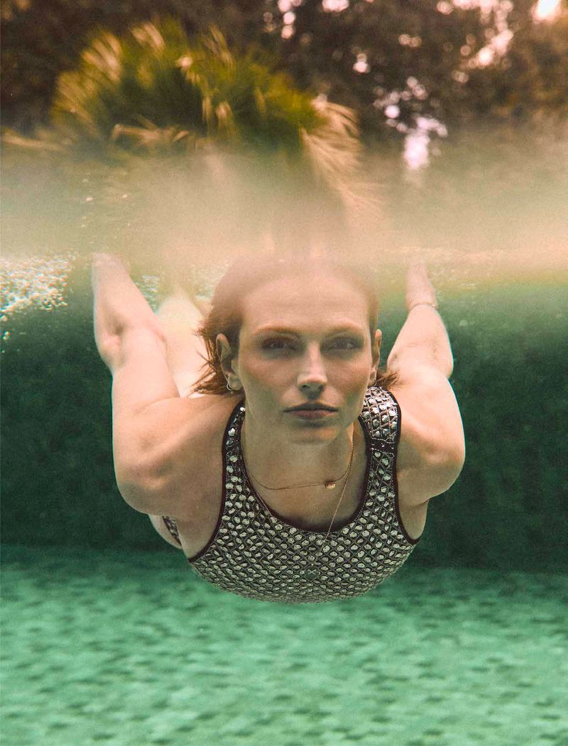 Underwater view of a woman with red hair wearing a patterned swimsuit. Bubbles and a blurred green background are visible.