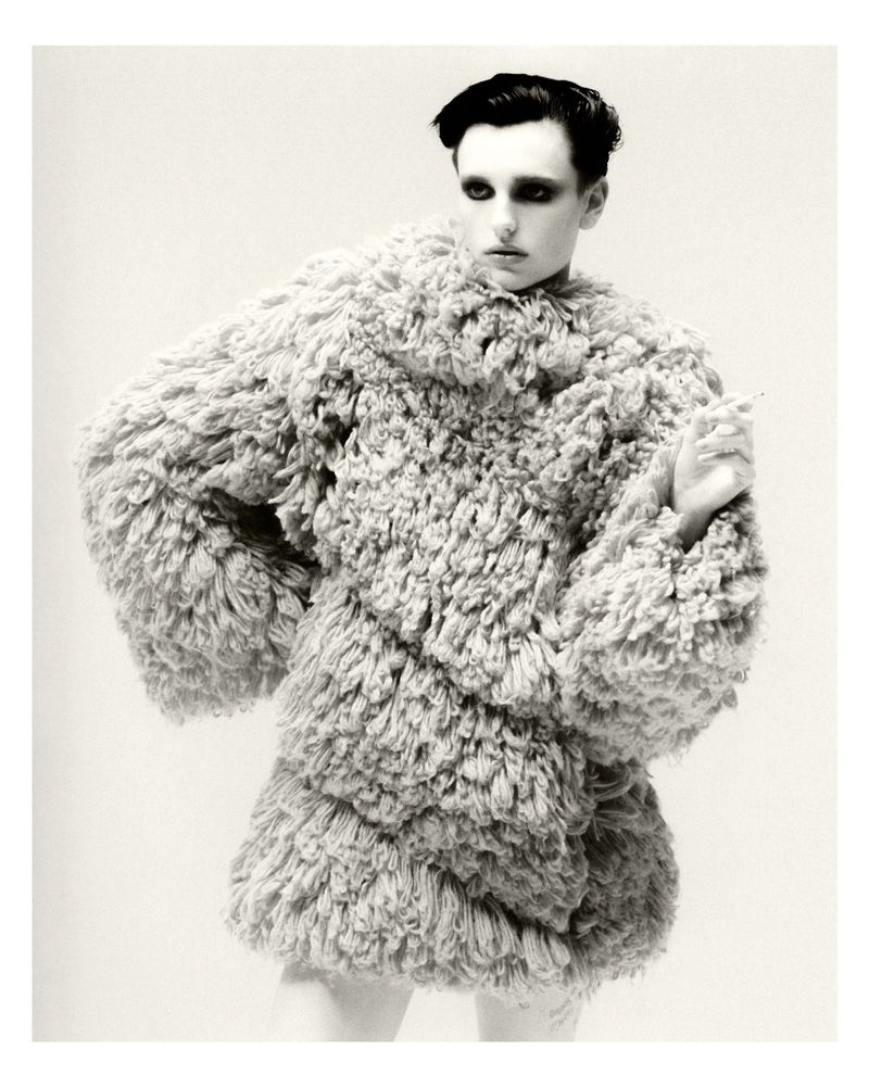 Studio portrait of an adult male fashion model wearing a large furry coat against a neutral background, with dark hair and dramatic makeup.