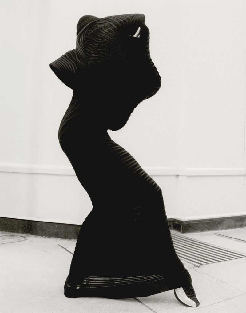 Person in a long, dark dress strikes a pose in a minimalist setting. The dress features a layered, ridged pattern. Studio shot.