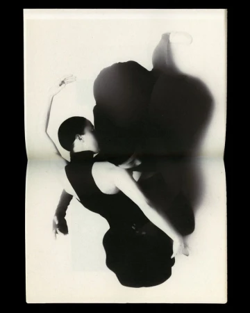 Black-and-white photograph features a woman in a dark dress striking a ballet pose against a light backdrop with a distinct shadow.