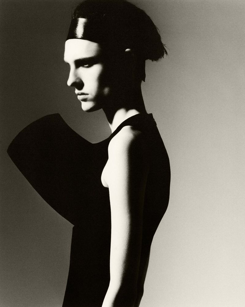 Studio portrait of a slender adult with dark hair and black geometric clothing against a neutral backdrop in black and white.