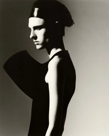 Studio portrait of a slender adult with dark hair and black geometric clothing against a neutral backdrop in black and white.