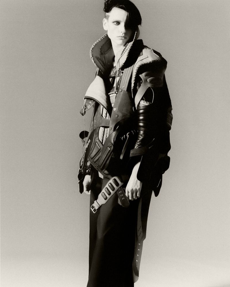 A person with pale skin and dark eye makeup stands in a studio, wearing a black beret, a leather jacket, and a long black skirt.