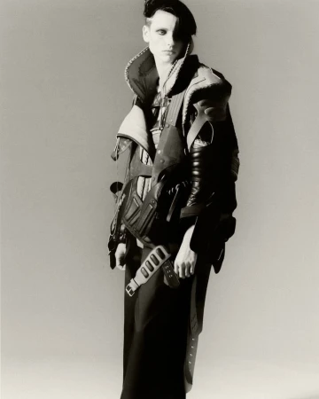 A person with pale skin and dark eye makeup stands in a studio, wearing a black beret, a leather jacket, and a long black skirt.