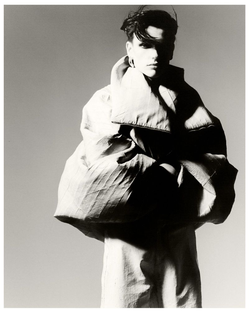 Monochrome fashion shot of a person with short dark hair, wearing an oversized light coat against a grey backdrop in a photo studio.