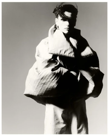 Monochrome fashion shot of a person with short dark hair, wearing an oversized light coat against a grey backdrop in a photo studio.