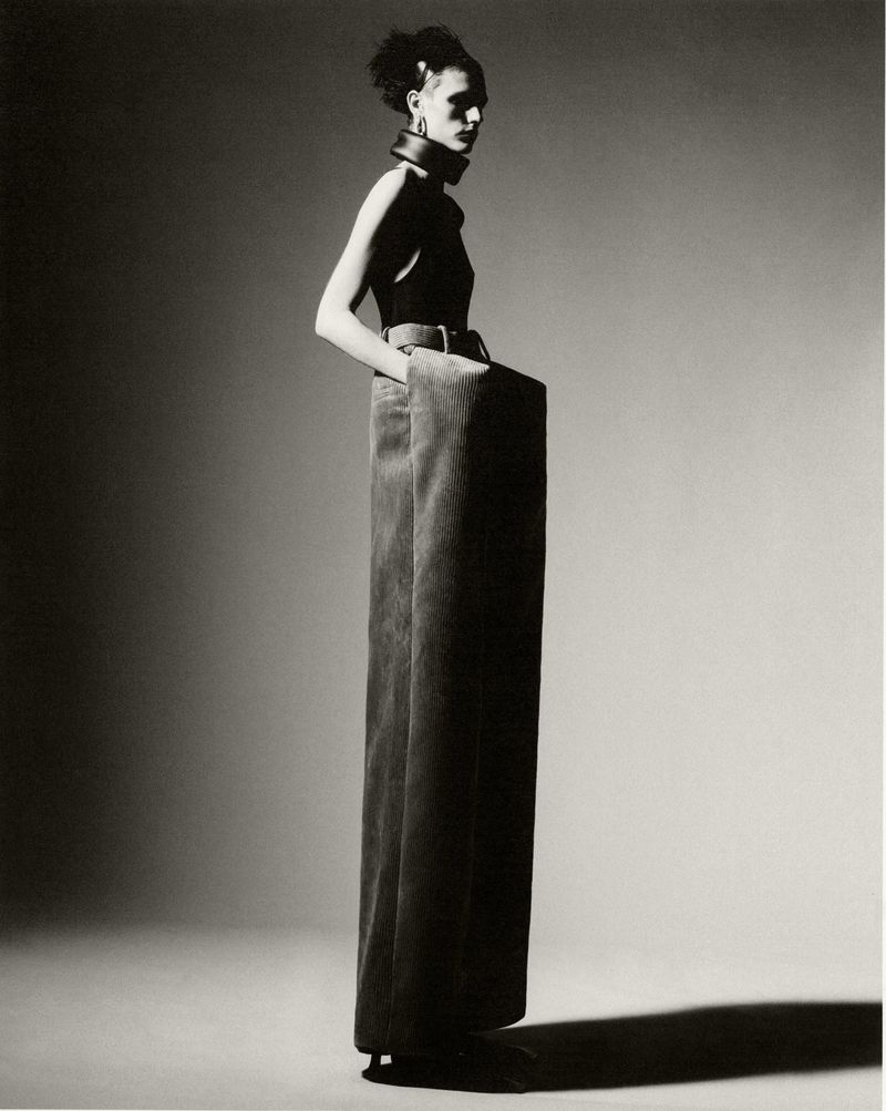 A woman poses in a sleeveless top and extra-wide-legged pants against a gradient gray background in a high-fashion studio shot.