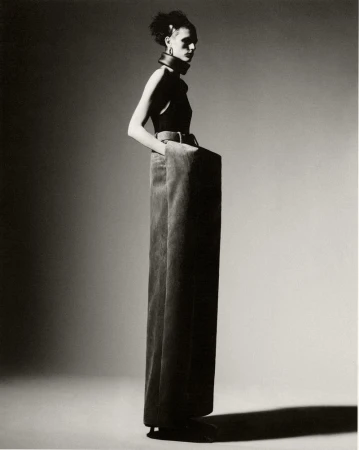 A woman poses in a sleeveless top and extra-wide-legged pants against a gradient gray background in a high-fashion studio shot.