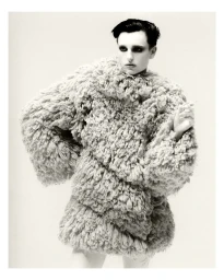 Model with dark styled hair wearing an oversized textured shaggy coat against a plain background