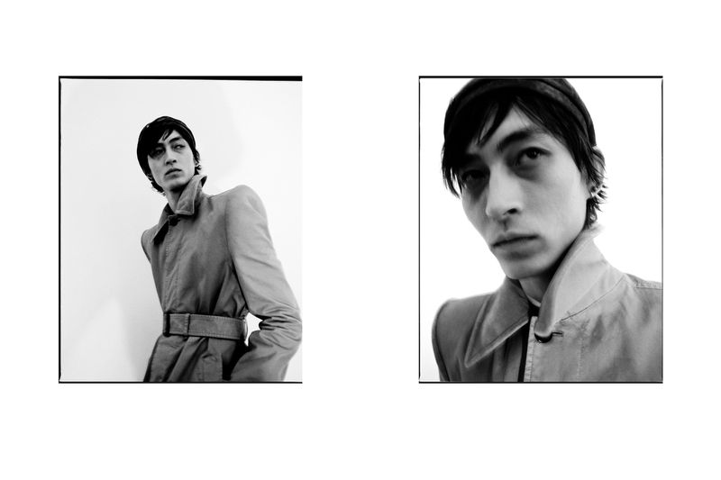 Black and white fashion portrait of a slender adult male with a trench coat. Studio lighting and a clean backdrop.