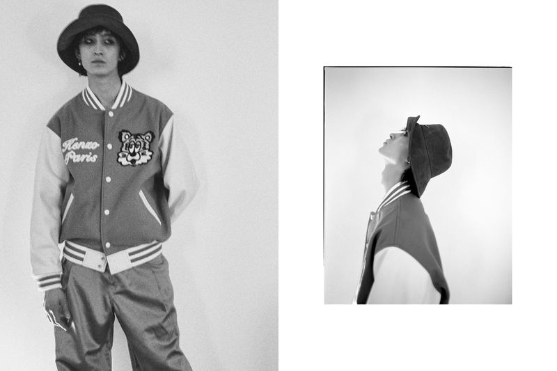 Black and white fashion shot of a young adult wearing a baseball jacket with a tiger patch and a bucket hat, standing in front of a white wall.