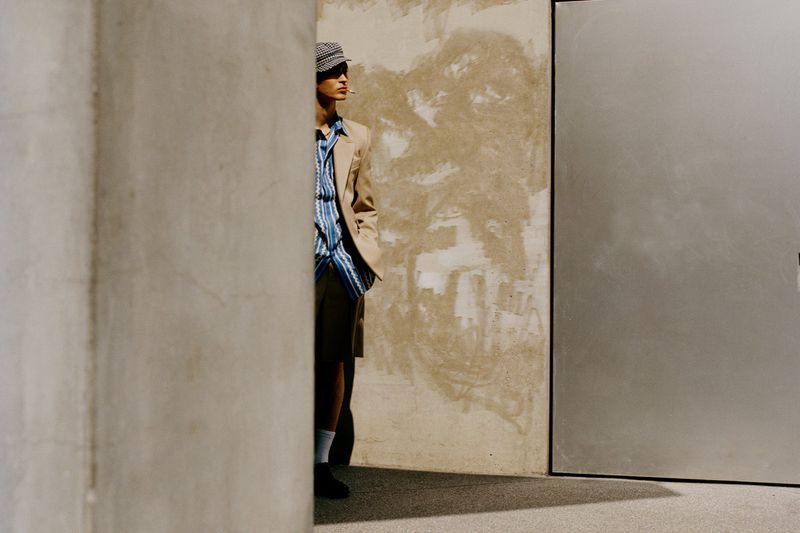 Adult male poses outdoors in a tan blazer, striped button-down, and cap with a cigarette in his mouth, standing against a textured wall.