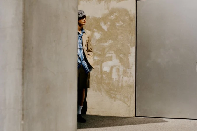 Adult male poses outdoors in a tan blazer, striped button-down, and cap with a cigarette in his mouth, standing against a textured wall.