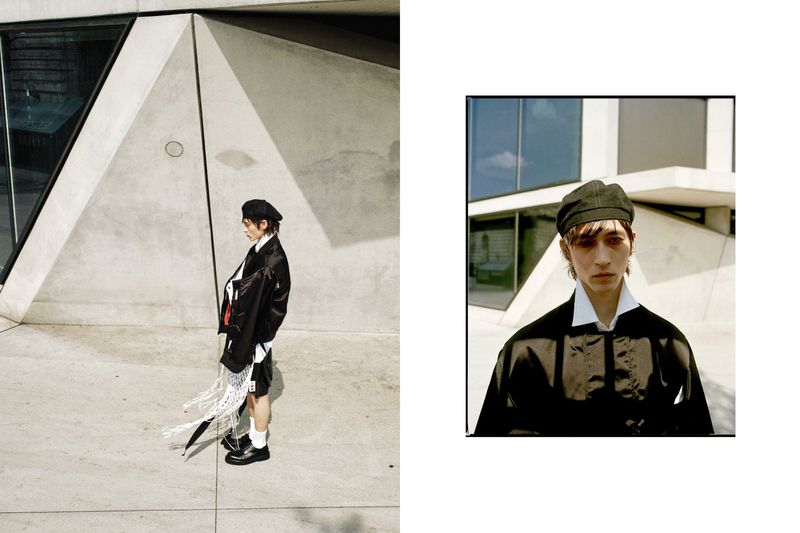 Fashion editorial shot of a young adult man in a black jacket and beret standing outside a modern building on a sunny day.