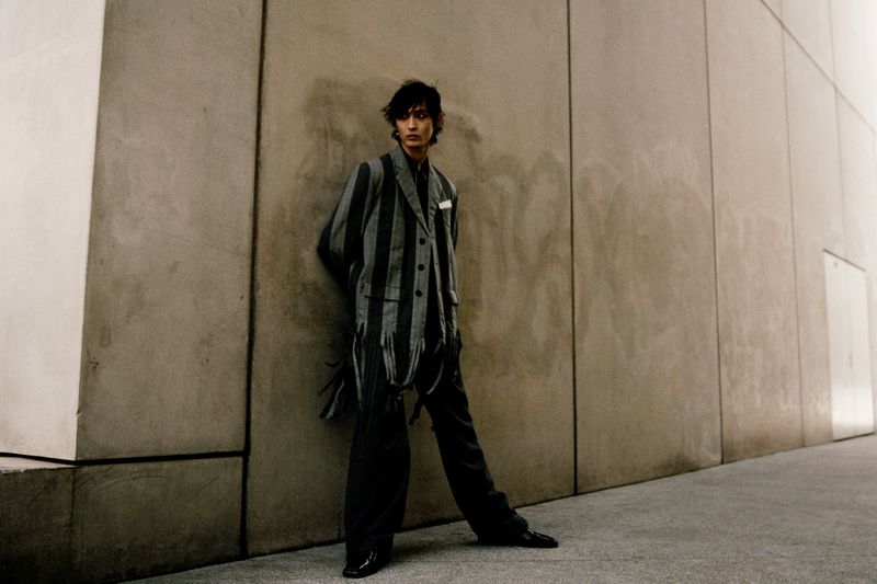 Adult male fashion model poses against a textured concrete wall wearing a gray striped blazer suit with a white pocket square.