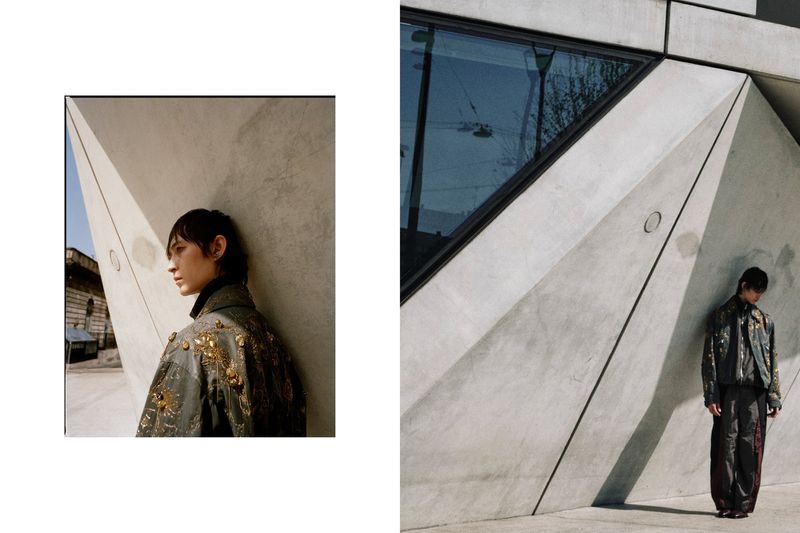 Editorial fashion shot of a person in an ornate jacket, posing against a geometrically complex concrete architectural structure.