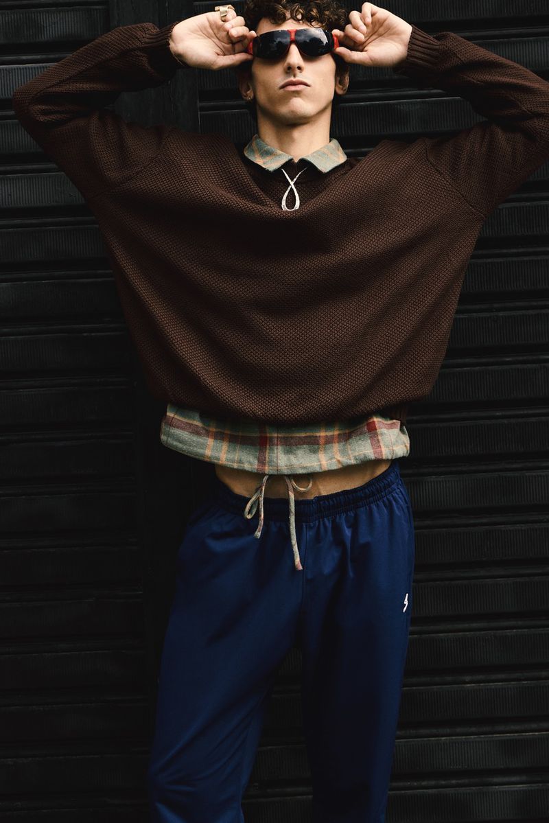 Stylish young man in brown sweater poses against a black background, accessorized with sunglasses, necklace, and blue pants.