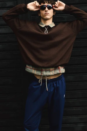 Stylish young man in brown sweater poses against a black background, accessorized with sunglasses, necklace, and blue pants.