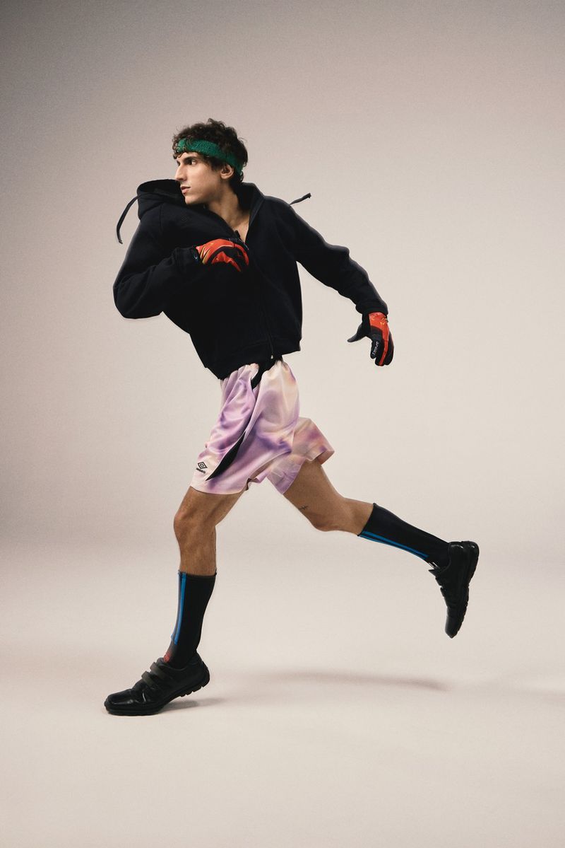 Athletic man in motion wearing black hoodie, pink shorts, gloves, shoes, and a headband against a neutral background in a studio setting.
