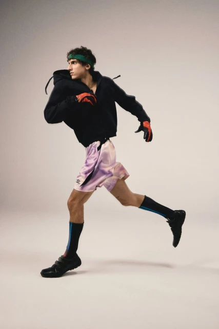 Athletic man in motion wearing black hoodie, pink shorts, gloves, shoes, and a headband against a neutral background in a studio setting.