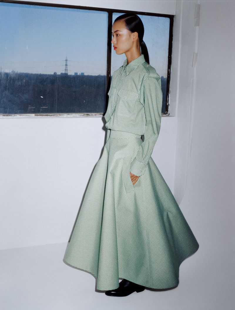 Asian woman in green gingham skirt and shirt stands in front of window with cityscape view. Fashion editorial shot, indoor setting.