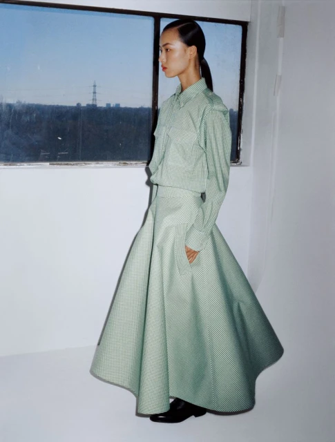 Asian woman in green gingham skirt and shirt stands in front of window with cityscape view. Fashion editorial shot, indoor setting.