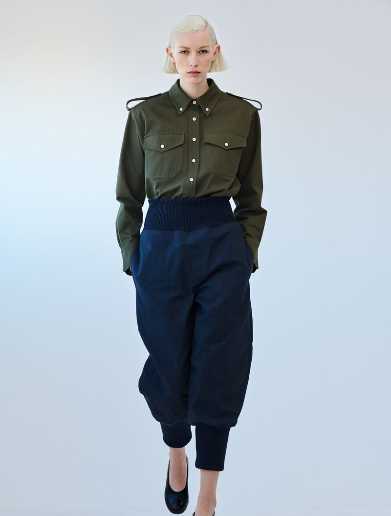 Fashion shot: woman with platinum blonde hair wearing an olive green button-up shirt, blue high-waisted pants, and black shoes on white.