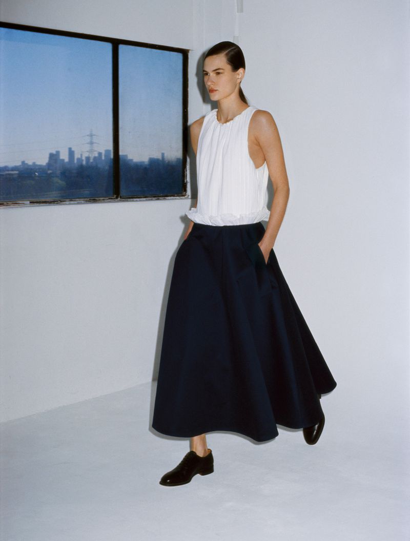 A slender woman poses in a sleeveless white blouse and a-line navy skirt against a white wall with a city skyline view.
