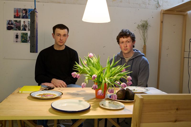 Two men pose at a wooden table with a vase of tulips, plates, and avocados in a brightly lit indoor setting with minimalist decor.