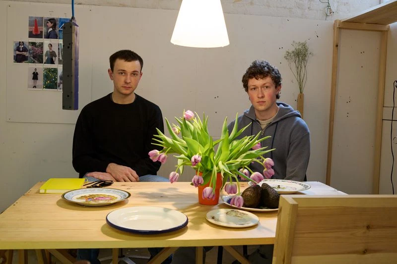 Two men pose at a wooden table with a vase of tulips, plates, and avocados in a brightly lit indoor setting with minimalist decor.