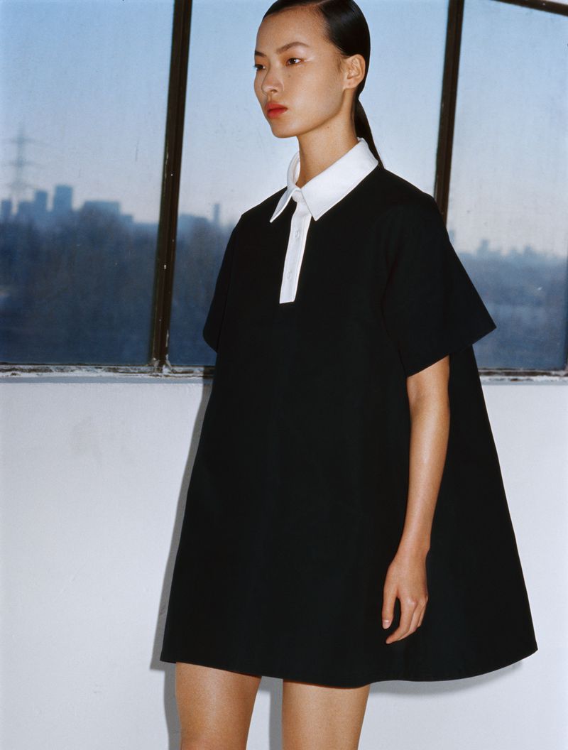 Asian woman in a black short dress with a white collar poses in a studio with a cityscape visible through a window. Fashion photography.