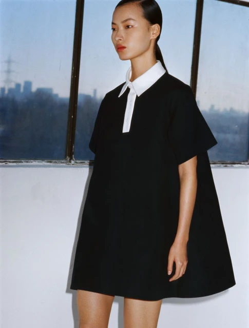 Asian woman in a black short dress with a white collar poses in a studio with a cityscape visible through a window. Fashion photography.