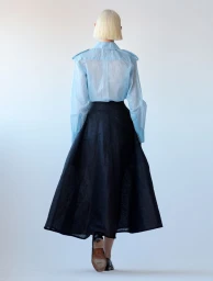 Model with blonde bob seen from behind wearing a light blue shirt and dark skirt against a plain backdrop.
