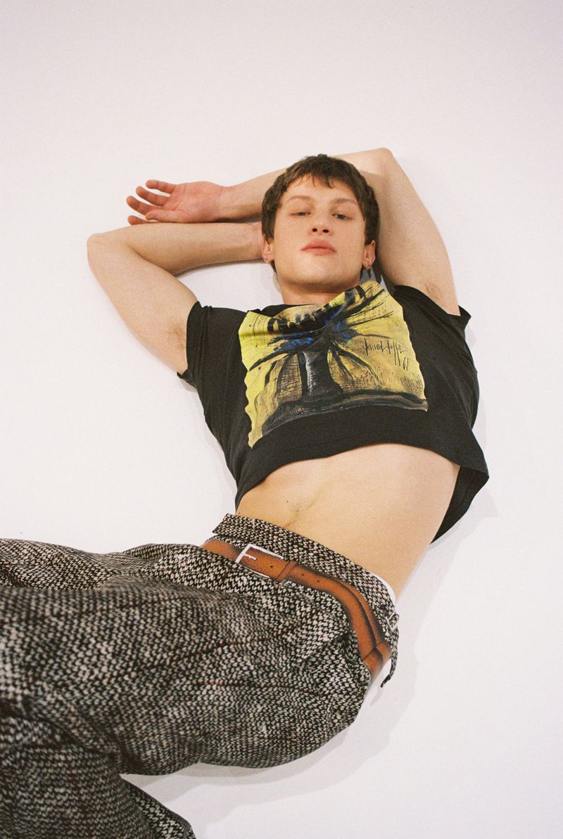 An adult male model poses while lying on a white surface wearing a graphic t-shirt, patterned pants, and brown belt in a studio.
