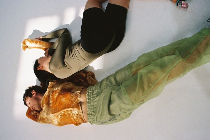 Two people, one in a green tracksuit and one in a spandex suit, laying on a white surface, in a fashion forward, editorial style.