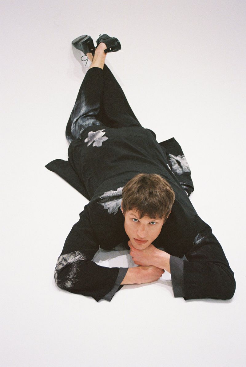 A young adult male lies on his stomach on a white surface wearing a floral print matching robe and pants with black dress shoes.