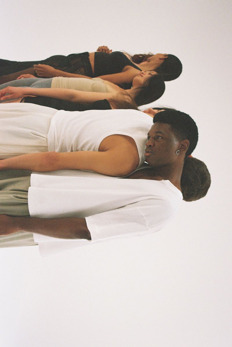 Group of diverse young adults lying head-to-toe in a row in bright studio lighting, wearing casual tops, against a neutral background.