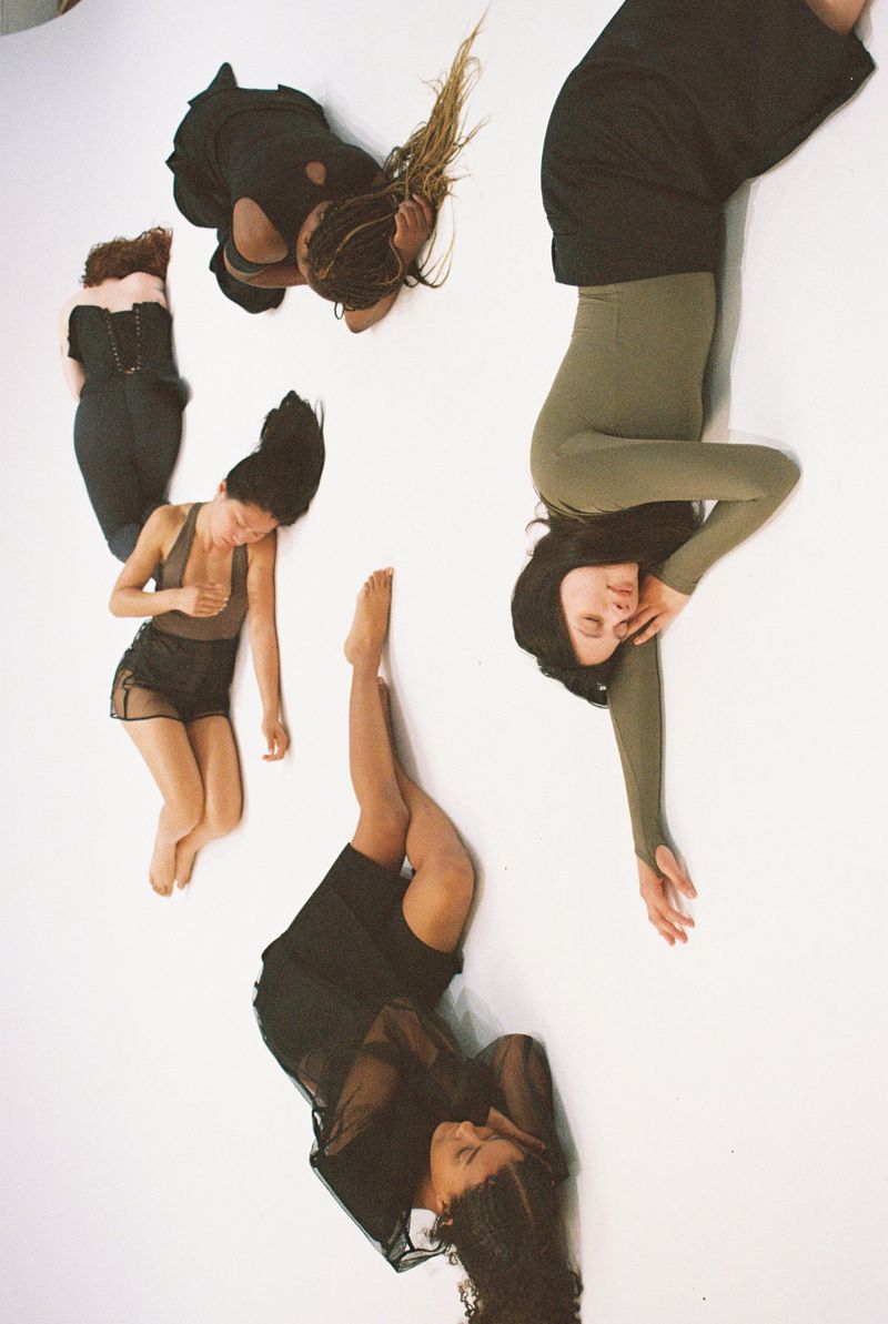 Overhead shot of five women lying on a white surface, wearing dark clothing and striking various poses, creating a visually artistic layout.