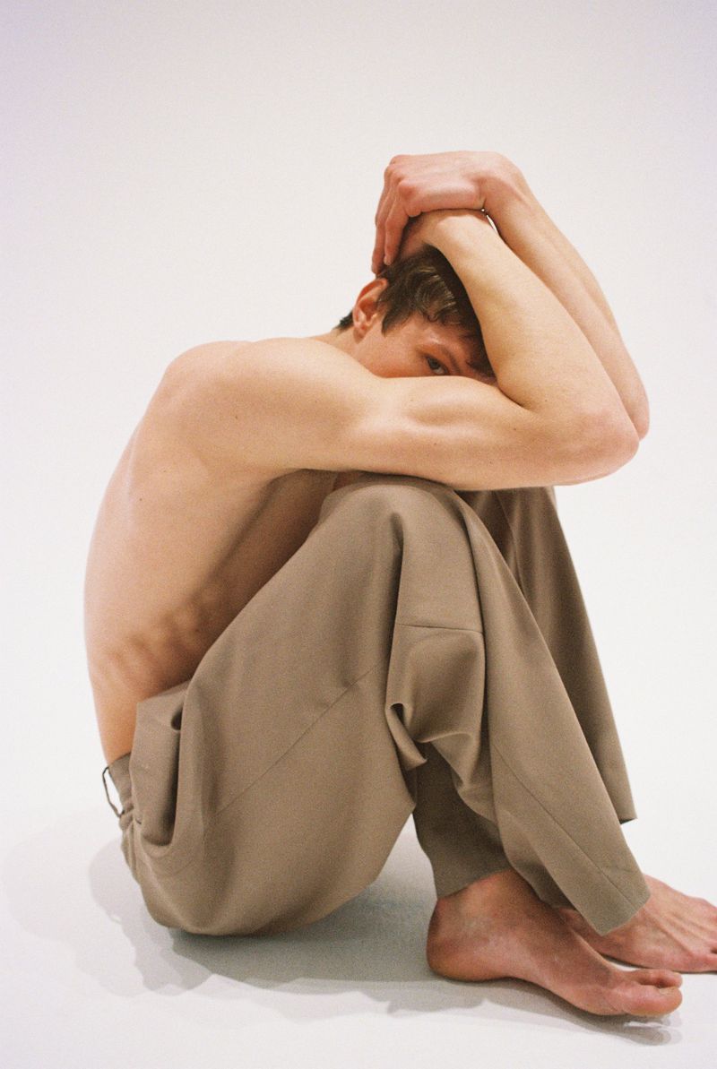 Young man sits with arms around legs in photo studio, wearing beige pants. Light skin, light background. Studio lighting shows muscles.
