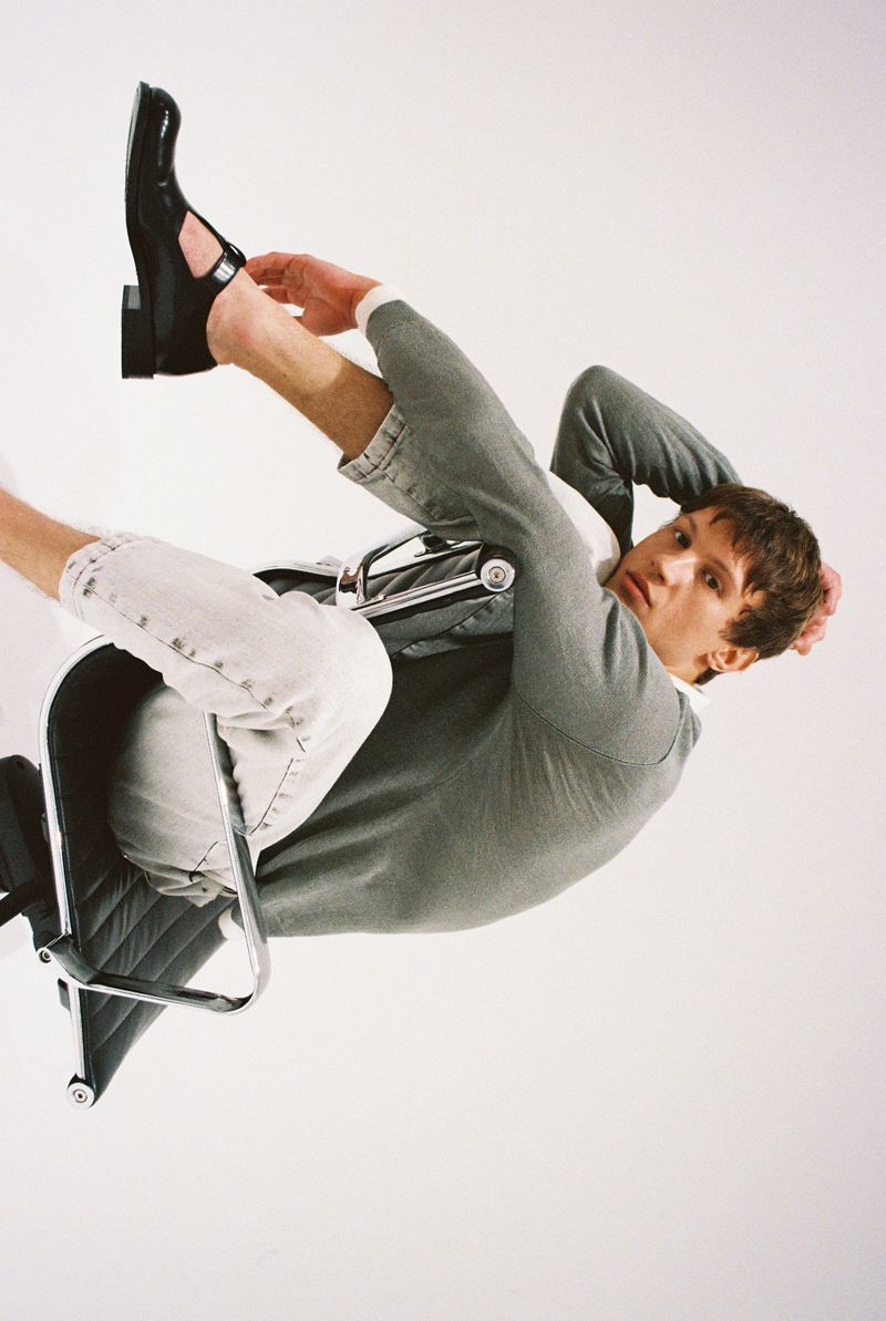 Adult male model in a grey long-sleeve shirt and light-wash jeans sits backward in a black office chair against a white backdrop.