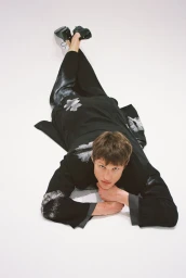 Model lying prone on a plain white studio background wearing a black patterned coat and trousers with black shoes.