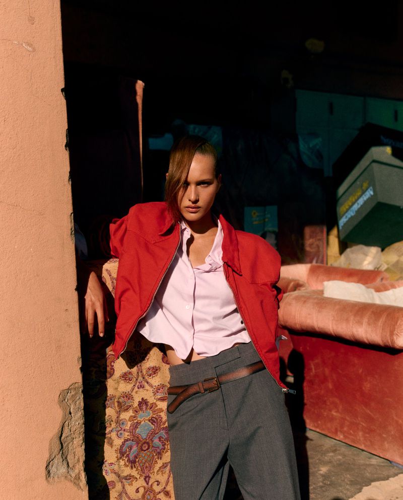Fashion shot of a woman in a red jacket and gray pants leaning against a wall, with a colorful couch and stacked boxes behind her.
