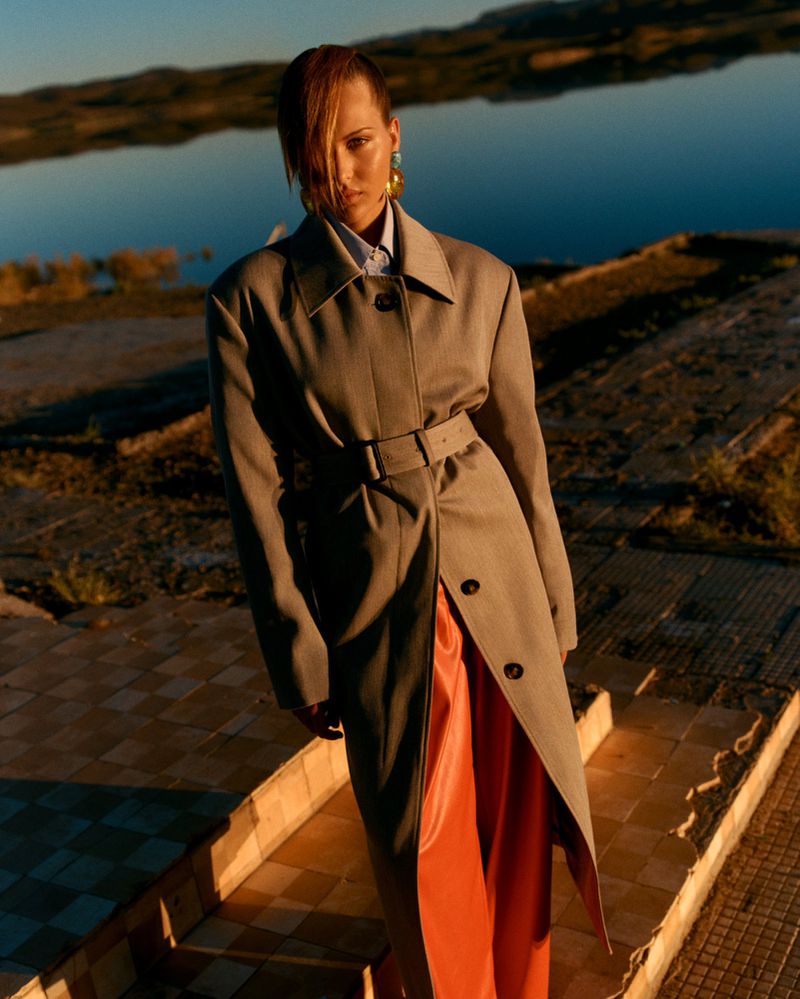 Woman in belted grey coat and orange dress poses on tiled terrace by reflective lake, bathed in warm sunlight, fashion editorial.