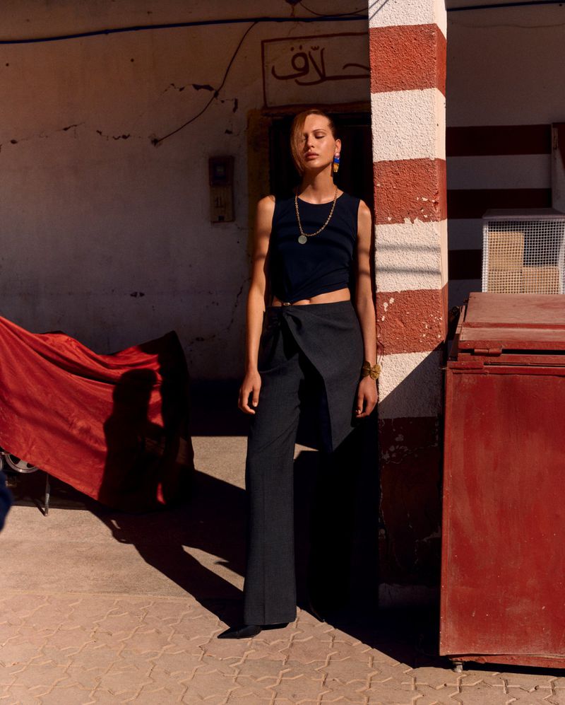An adult female model poses in black crop top and dark pants against a textured exterior with warm lighting, red drapery, and columns.