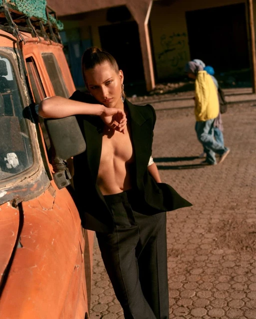 Woman in a black blazer leans against an orange vintage car in a desert setting with two children walking in the background.