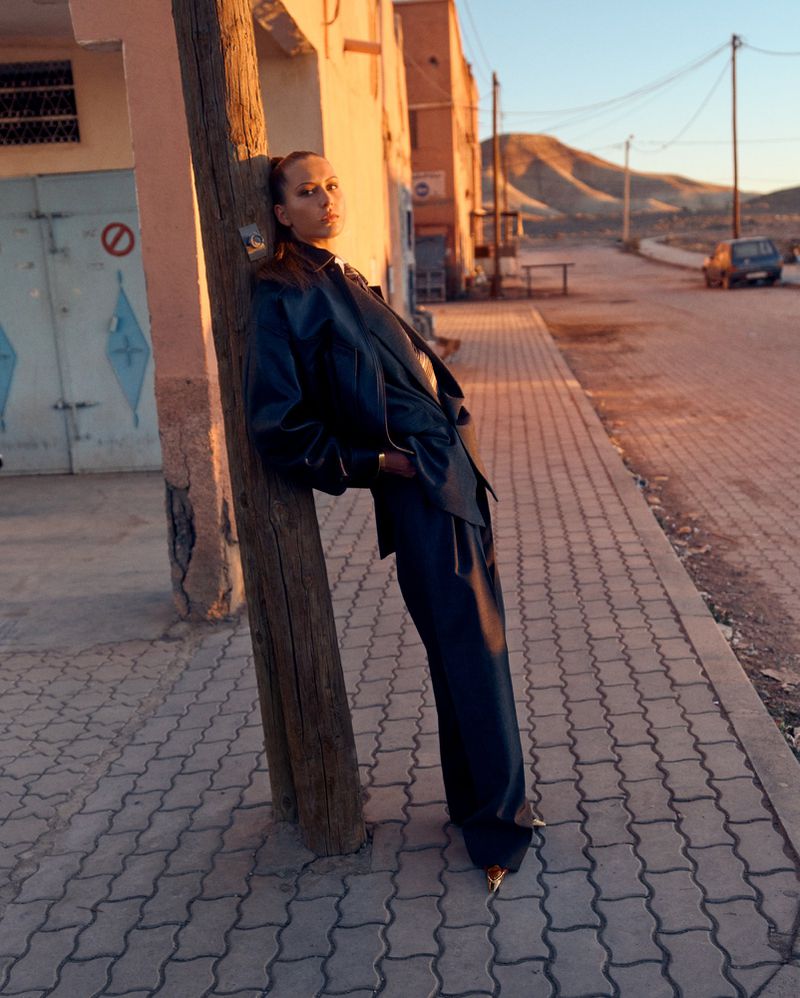 Woman in dark suit and golden shoes leans on wooden pole in street in outdoor fashion shot with buildings and hills in the background.