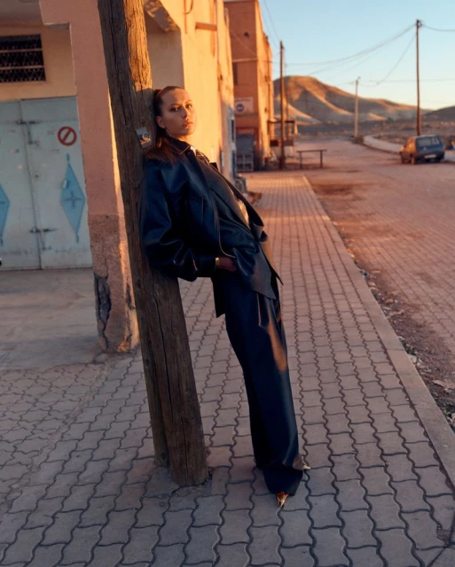 Woman in dark suit and golden shoes leans on wooden pole in street in outdoor fashion shot with buildings and hills in the background.