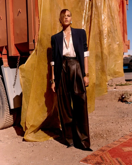 Fashion model poses in oversized blazer and wide-leg trousers next to truck and yellow plastic tarp in an outdoor setting.