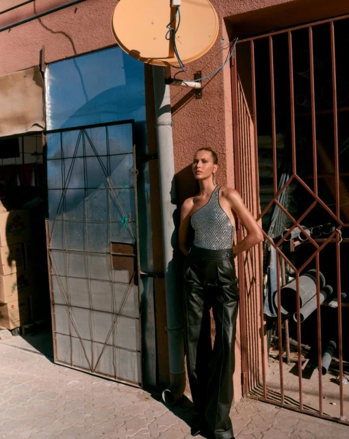 Fashion shot of a model in a one-shoulder metallic top and black trousers, posing against a weathered building with a satellite dish.