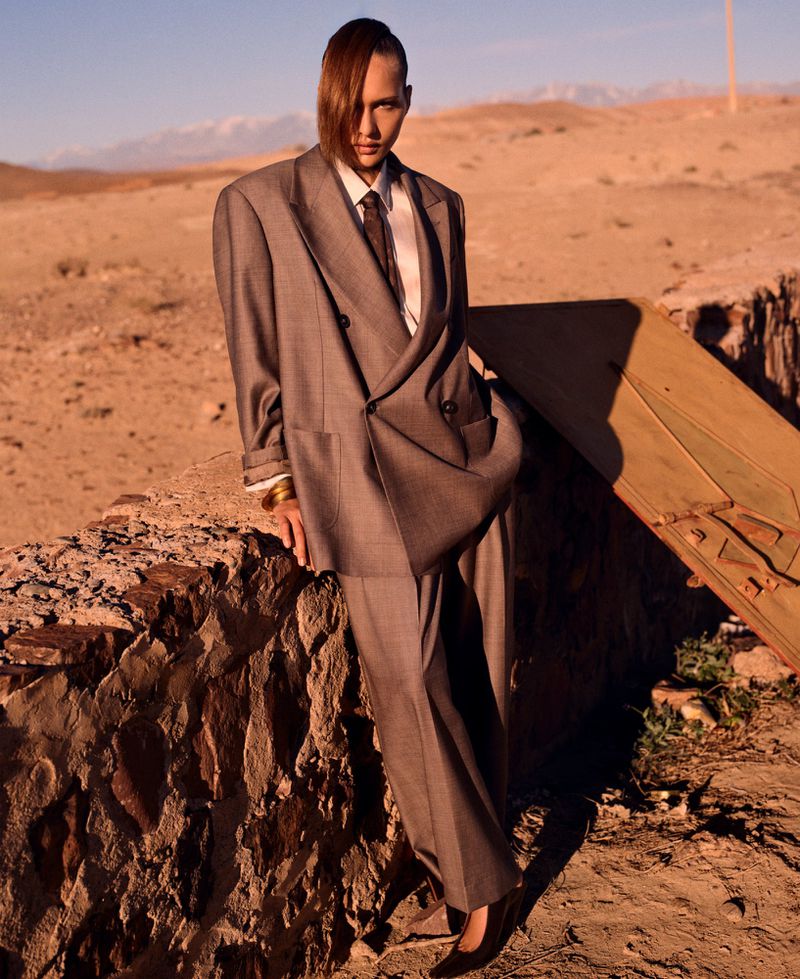 A person in a grey oversized suit poses in a desert landscape with rocky foreground, distant mountains, and a somber, fashionable aesthetic.